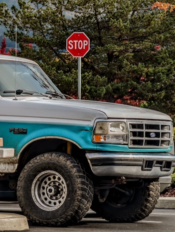 A teal and silver Ford F-250 truck is parked next to a stop sign. The truck appears rugged with large off-road tires and is positioned in a natural setting with dense green foliage and some red leaves in the background.