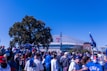 A large crowd of people, many wearing blue and red clothing, gathered outside a sports stadium with a large 'T' logo and signage for 'Globe Life Field' visible. An array of merchandise including baseball caps and jerseys can be seen. In the background, a large tree and clear blue sky are visible.