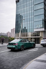 A green sports car is parked on a modern urban street in front of a tall glass building. A person is walking on the sidewalk, and another white vehicle is parked nearby. The environment appears clean and sophisticated, with architectural elements emphasizing vertical lines.