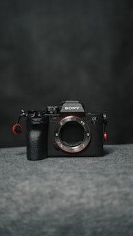 A dark-toned digital camera body with prominent branding and controls, placed on a soft, textured surface.