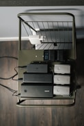 A metal cart holds several electronic charging devices and batteries neatly arranged on a shelf. The cart is on a wooden floor, and there's a wall socket visible with connected plugs.