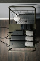 A metal cart holds several electronic charging devices and batteries neatly arranged on a shelf. The cart is on a wooden floor, and there's a wall socket visible with connected plugs.