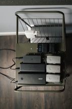 A metal cart holds several electronic charging devices and batteries neatly arranged on a shelf. The cart is on a wooden floor, and there's a wall socket visible with connected plugs.