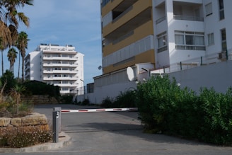 A residential area featuring multi-story apartment buildings with balconies. A barrier gate is positioned in the driveway, partially obstructed by surrounding foliage. Palm trees and shrubs are visible, offering a contrast to the urban setting. The sky is clear and blue.