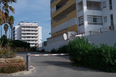 A residential area featuring multi-story apartment buildings with balconies. A barrier gate is positioned in the driveway, partially obstructed by surrounding foliage. Palm trees and shrubs are visible, offering a contrast to the urban setting. The sky is clear and blue.