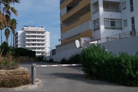 A residential area featuring multi-story apartment buildings with balconies. A barrier gate is positioned in the driveway, partially obstructed by surrounding foliage. Palm trees and shrubs are visible, offering a contrast to the urban setting. The sky is clear and blue.