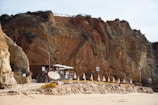 Wooden hut and umbrellas are situated on a sandy beach against a backdrop of large, rugged cliffs. The structure appears to be a small beach bar or café with tables and chairs under shade. White umbrellas are lined up in a row alongside sunbeds, suggesting a leisure area.