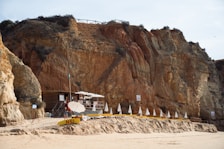Wooden hut and umbrellas are situated on a sandy beach against a backdrop of large, rugged cliffs. The structure appears to be a small beach bar or café with tables and chairs under shade. White umbrellas are lined up in a row alongside sunbeds, suggesting a leisure area.