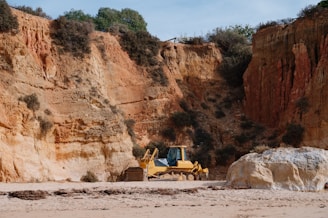 A construction vehicle is stationed on a sandy beach, adjacent to towering rocky cliffs with patches of vegetation. The cliffs display layers of rich, earthy tones ranging from sandy beige to deeper ochre, creating a warm contrast against the clear sky above.