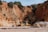 A construction vehicle is stationed on a sandy beach, adjacent to towering rocky cliffs with patches of vegetation. The cliffs display layers of rich, earthy tones ranging from sandy beige to deeper ochre, creating a warm contrast against the clear sky above.