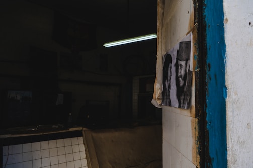A dimly lit room with a partially visible poster on a tiled wall. The poster features two individuals who appear to be smiling. The room has a worn-out look with dark shadows and a single fluorescent light illuminating part of the scene. The outer tiles have chipping paint, revealing a rustic interior.