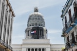 A grand dome structure is framed by two older buildings on either side. The dome is covered in scaffolding, indicating renovations. A Cuban flag is prominently displayed, suggesting the location is in Cuba.