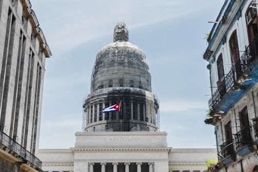 A grand dome structure is framed by two older buildings on either side. The dome is covered in scaffolding, indicating renovations. A Cuban flag is prominently displayed, suggesting the location is in Cuba.