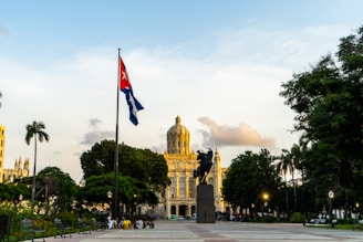 A plaza with a large statue in the center, surrounded by trees and benches. A prominent building with a dome is visible in the background. A flagpole with the Cuban flag is on the left side of the image. People are gathered near the statue, and the atmosphere is calm with a clear sky overhead.