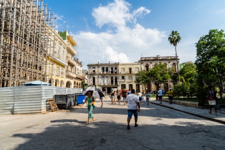 A street scene with people walking along a cobblestone road, surrounded by a mix of colonial-style buildings and modern construction scaffolding. The sky is clear with some clouds, and lush greenery is visible, including a tall palm tree. The atmosphere appears lively with several pedestrians enjoying the sunny weather, and a few using umbrellas for shade.
