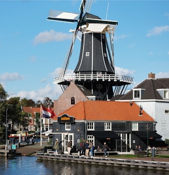 A traditional Dutch windmill stands prominently against a clear blue sky, surrounded by charming red-roofed brick buildings. The scene is set by a calm canal, with people casually gathering near the water. A Dutch flag is visible, adding a touch of national pride to the picturesque landscape.