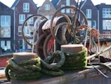 A collection of metal gears and pulleys intertwined with ropes on a boat. The boat appears weathered with rust visible on the machinery, and moss-covered ropes wound around wooden posts. In the background, there are several multi-story brick buildings with large windows.
