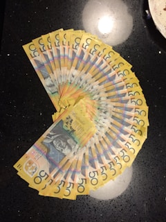 A fan-like arrangement of multiple Australian fifty-dollar notes is spread out on a dark, reflective surface. The notes display intricate designs and a portrait along with vibrant colors.