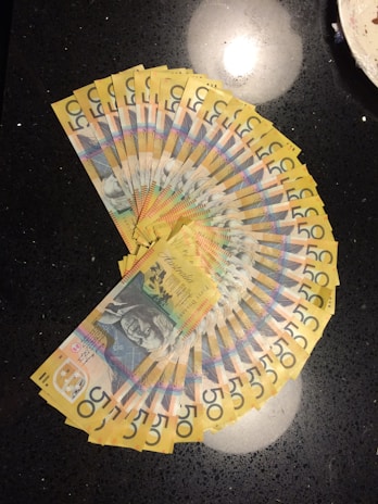 A fan-like arrangement of multiple Australian fifty-dollar notes is spread out on a dark, reflective surface. The notes display intricate designs and a portrait along with vibrant colors.