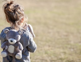 A child with their back turned, wearing a gray hoodie and a backpack designed like a stuffed teddy bear. The child holds a soft bunny toy. The background is a blurred, grassy field.