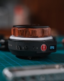 A close-up of a small device sits on a surface, featuring a wooden texture on the top and a black base with several buttons including one labeled 'REC' in red. There is also a small digital display showing the number five, and the underlying surface has a green grid pattern.