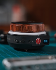 A close-up of a small device sits on a surface, featuring a wooden texture on the top and a black base with several buttons including one labeled 'REC' in red. There is also a small digital display showing the number five, and the underlying surface has a green grid pattern.