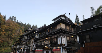 A traditional Japanese building, possibly an inn or a ryokan, is situated in a forested area with dense trees in the background. The architecture features wooden elements, multiple levels with balconies, and a prominent sign with Japanese characters. The setting appears serene and picturesque, evoking a sense of tranquility.