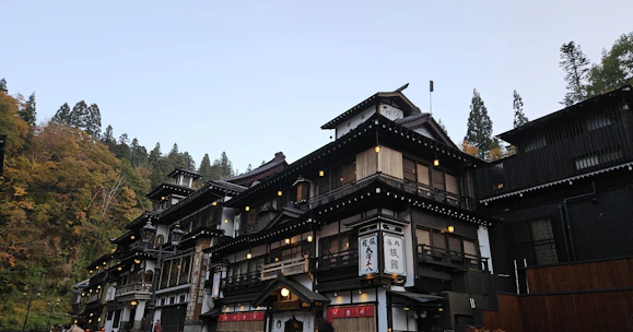 A traditional Japanese building, possibly an inn or a ryokan, is situated in a forested area with dense trees in the background. The architecture features wooden elements, multiple levels with balconies, and a prominent sign with Japanese characters. The setting appears serene and picturesque, evoking a sense of tranquility.