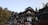 A traditional Japanese building, possibly an inn or a ryokan, is situated in a forested area with dense trees in the background. The architecture features wooden elements, multiple levels with balconies, and a prominent sign with Japanese characters. The setting appears serene and picturesque, evoking a sense of tranquility.