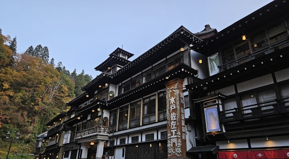 A traditional multi-story Japanese building features intricate wooden architecture and ornate details. The structure is surrounded by lush greenery with trees showcasing autumn foliage. The building's facade is adorned with lanterns and a prominent vertical sign with Japanese characters. The sky above is clear, providing a serene backdrop.