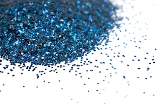 A collection of small, square blue glitter particles spread over a white surface, with many particles densely gathered and some scattered sparsely, reflecting light.