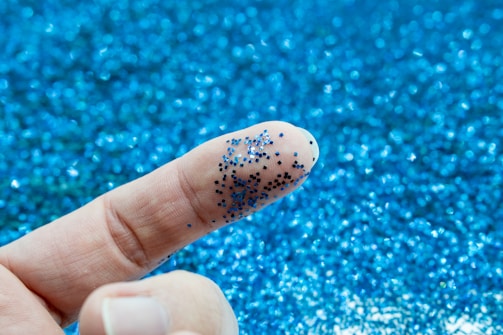 A close-up of a finger covered in small blue glitter particles, set against a background filled with more scattered blue glitter.