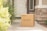 A cardboard box labeled 'MealPro' is placed on a porch next to some grass and a brick wall. The porch leads to a white door with a glass window panel. There are plants on one side and the entryway appears clean and brightly lit.