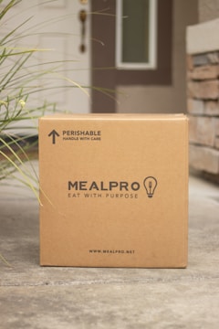 A cardboard box labeled 'MEALPRO' with the tagline 'EAT WITH PURPOSE' is placed outside a house. The box has a label indicating it contains perishable items and should be handled with care. There is grass visible to the left side and a brick wall partially visible on the right. The house door and part of a window can be seen in the background.