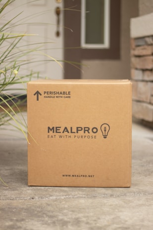 A cardboard box labeled 'MEALPRO' with the tagline 'EAT WITH PURPOSE' is placed outside a house. The box has a label indicating it contains perishable items and should be handled with care. There is grass visible to the left side and a brick wall partially visible on the right. The house door and part of a window can be seen in the background.