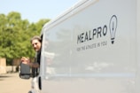 A man is leaning out of the driver's window of a white van, waving his hand. The side of the van is branded with the logo and slogan of MealPro, a company that provides meals, emphasizing fitness and athleticism. The background features greenery visible in soft focus.