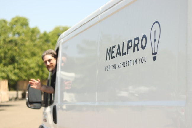 A man is leaning out of the driver's window of a white van, waving his hand. The side of the van is branded with the logo and slogan of MealPro, a company that provides meals, emphasizing fitness and athleticism. The background features greenery visible in soft focus.