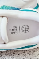 An image of a sneaker with a visible insole featuring the text 'END PLASTIC WASTE' alongside a globe symbol. The sneaker's interior includes shades of white and accents of teal. It is placed on a textured carpet surface.