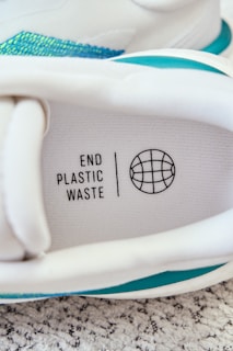 An image of a sneaker with a visible insole featuring the text 'END PLASTIC WASTE' alongside a globe symbol. The sneaker's interior includes shades of white and accents of teal. It is placed on a textured carpet surface.