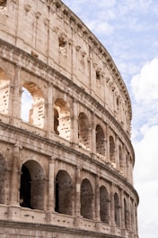 The image features a section of an ancient amphitheater with rows of arches and large stone blocks, showcasing Roman architectural elements. The sky in the background is partly cloudy, creating a dramatic contrast with the weathered stone facade.
