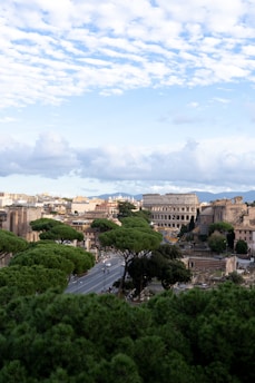 An expansive view of a historical cityscape featuring the Colosseum with numerous ancient structures amidst lush green trees. A wide road lined with trees runs through the scene, leading towards the iconic Roman architecture. The sky overhead is filled with scattered clouds, creating a bright and airy atmosphere.
