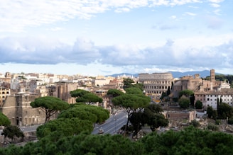 A panoramic view of a historic city with ancient Roman architecture. The iconic Colosseum stands prominently, surrounded by lush green trees and classical buildings. The sky is partly cloudy, providing a soft, tranquil backdrop.