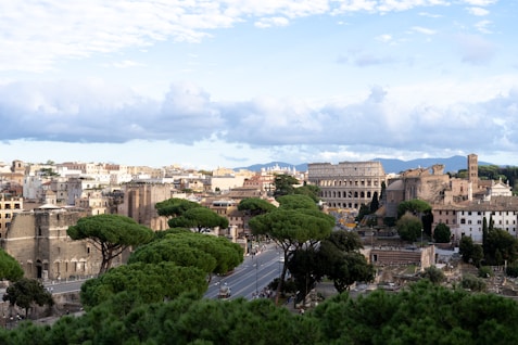 A panoramic view of a historic city with ancient Roman architecture. The iconic Colosseum stands prominently, surrounded by lush green trees and classical buildings. The sky is partly cloudy, providing a soft, tranquil backdrop.