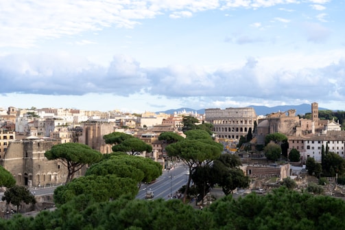 A panoramic view of a historic city with ancient Roman architecture. The iconic Colosseum stands prominently, surrounded by lush green trees and classical buildings. The sky is partly cloudy, providing a soft, tranquil backdrop.