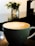 A cup of coffee featuring latte art in the shape of a leaf pattern sits on a dark reflective surface. In the background, there's a blurred view of greenery in a container, contributing to a serene atmosphere.