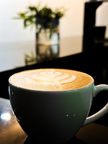 A cup of coffee featuring latte art in the shape of a leaf pattern sits on a dark reflective surface. In the background, there's a blurred view of greenery in a container, contributing to a serene atmosphere.
