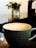 A cup of coffee featuring latte art in the shape of a leaf pattern sits on a dark reflective surface. In the background, there's a blurred view of greenery in a container, contributing to a serene atmosphere.