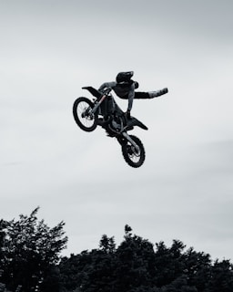 A motocross rider performs an aerial stunt mid-air, hanging off one side of the bike with both legs stretched out. The silhouette is set against a cloudy sky, and trees are visible at the bottom of the frame.