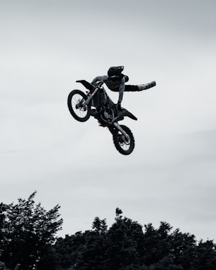 A motocross rider performs an aerial stunt mid-air, hanging off one side of the bike with both legs stretched out. The silhouette is set against a cloudy sky, and trees are visible at the bottom of the frame.