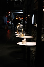 A dimly lit outdoor dining area on a city street, with several small round tables lined up. Each table has an unlit candle and upside-down glasses, suggesting a restaurant setup. The background shows blurred pedestrians and softly glowing city lights, creating a serene nighttime atmosphere.